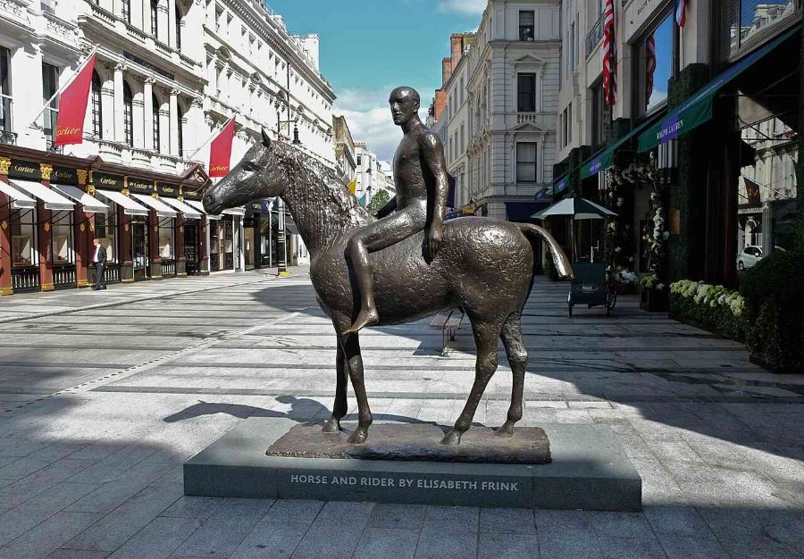 Bronze statue 'Horse and Rider' by Elisabeth Frink set on a pedestrian shopping street with branded storefronts and outdoor greenery.