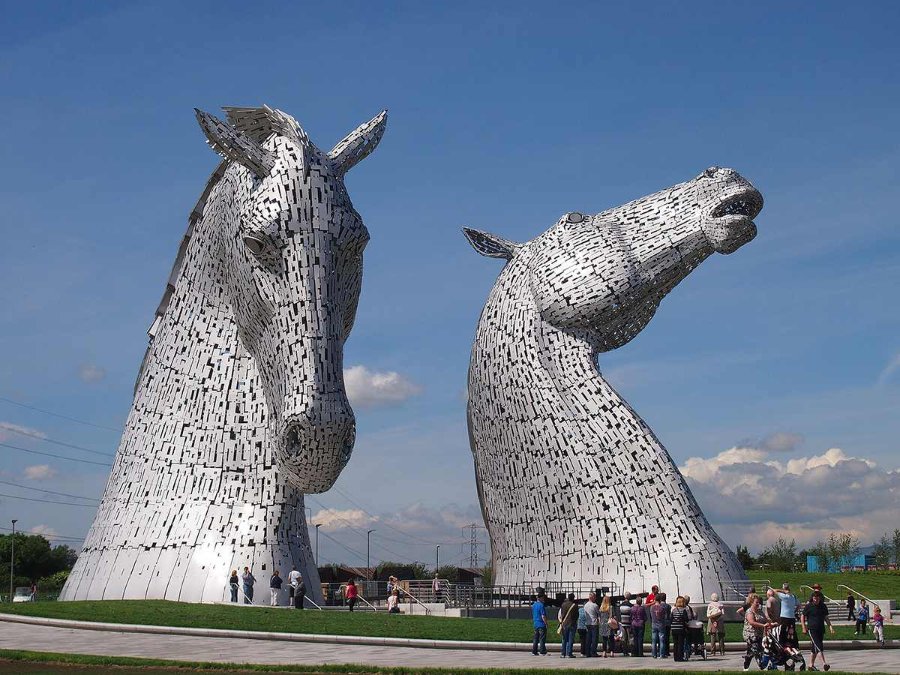 Two towering metallic horse head sculptures known as The Kelpies, surrounded by visitors and set against a clear blue sky.