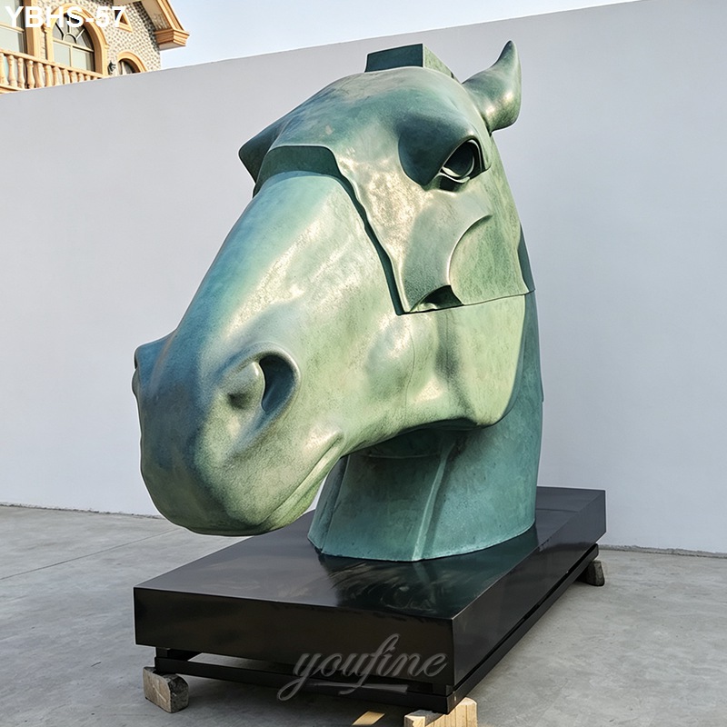 A large bronze horse head sculpture displayed on a black platform outdoors against a white wall with part of a building in the background.