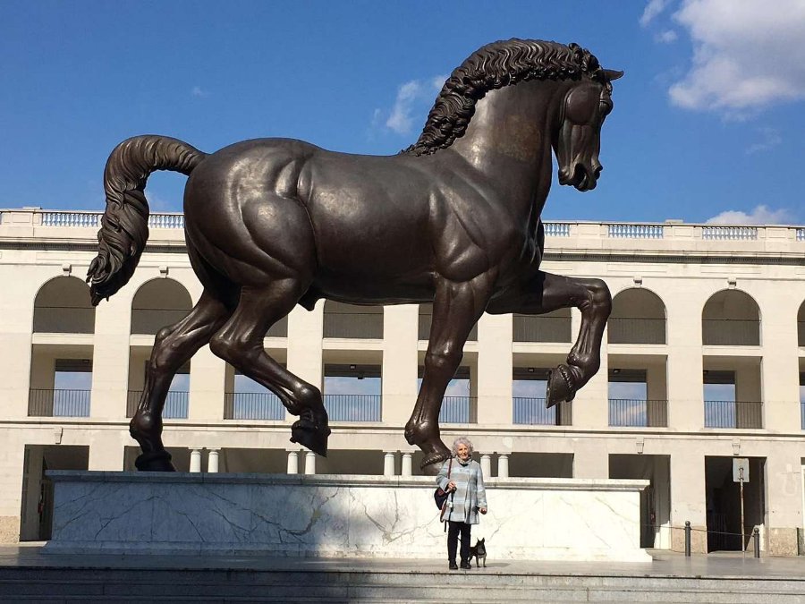 A massive bronze Leonardo's Horse sculpture on a marble pedestal in an open plaza, with a person standing nearby for scale under a bright sky.