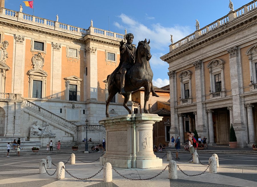 Bronze equestrian statue of Marcus Aurelius on a pedestal in Capitoline Hill, framed by historic buildings and tourist activity.