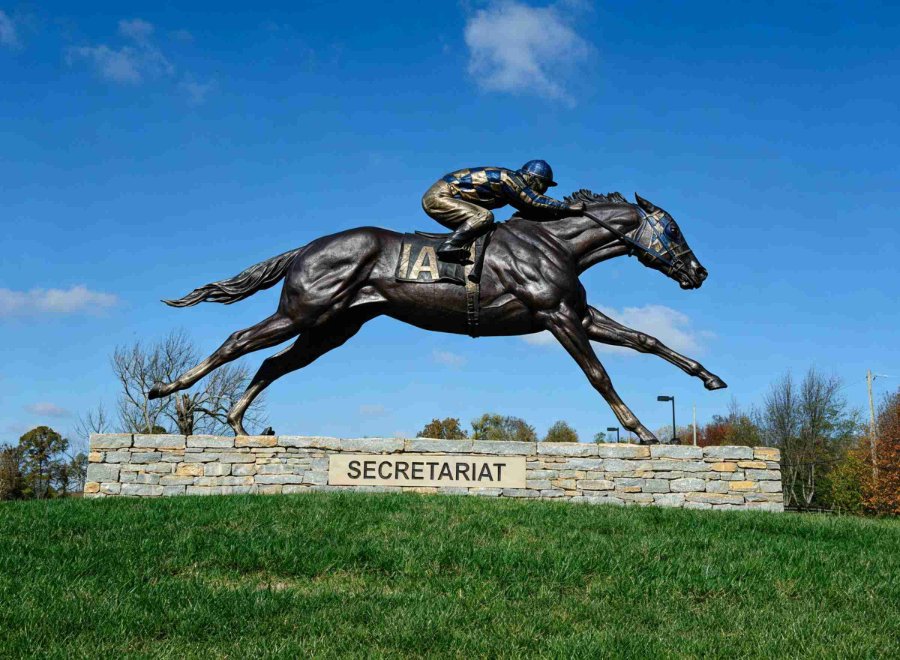 Bronze statue of Secretariat with jockey in racing position, mounted on a stone base labeled 'Secretariat,' surrounded by green grass and clear blue sky.