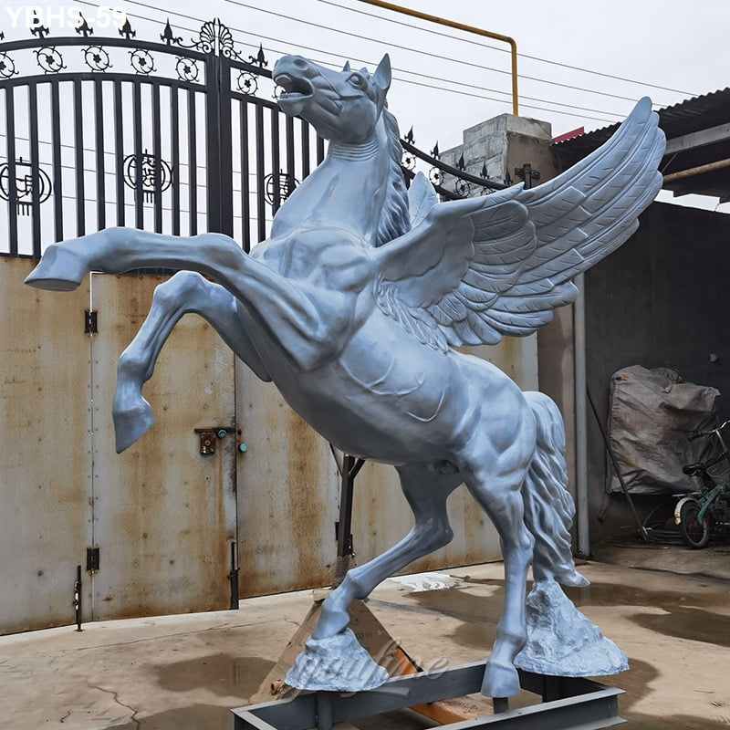 A winged horse bronze sculpture in a rearing position, captured outdoors near a decorative metal gate and industrial elements.