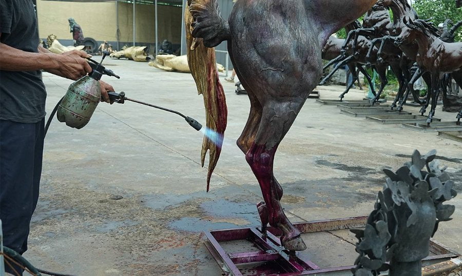 An artisan using heat and chemicals to apply patina to a bronze horse sculpture in an outdoor workshop setting.
