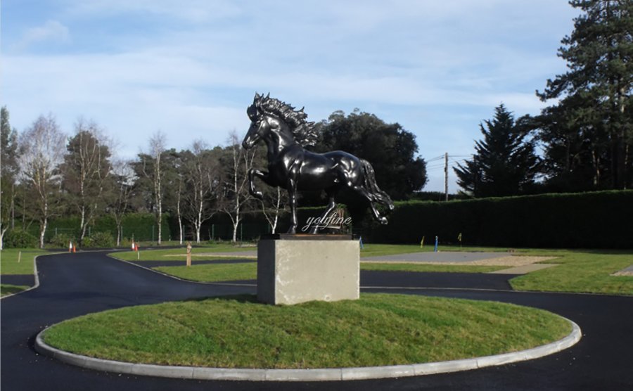A glossy black bronze horse statue displayed on a concrete pedestal in the center of a landscaped roundabout, surrounded by trees and grass.