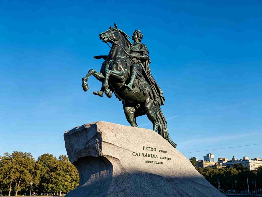 The Bronze Horseman statue of Peter the Great riding a rearing horse on a stone pedestal with blue sky and buildings in the background.