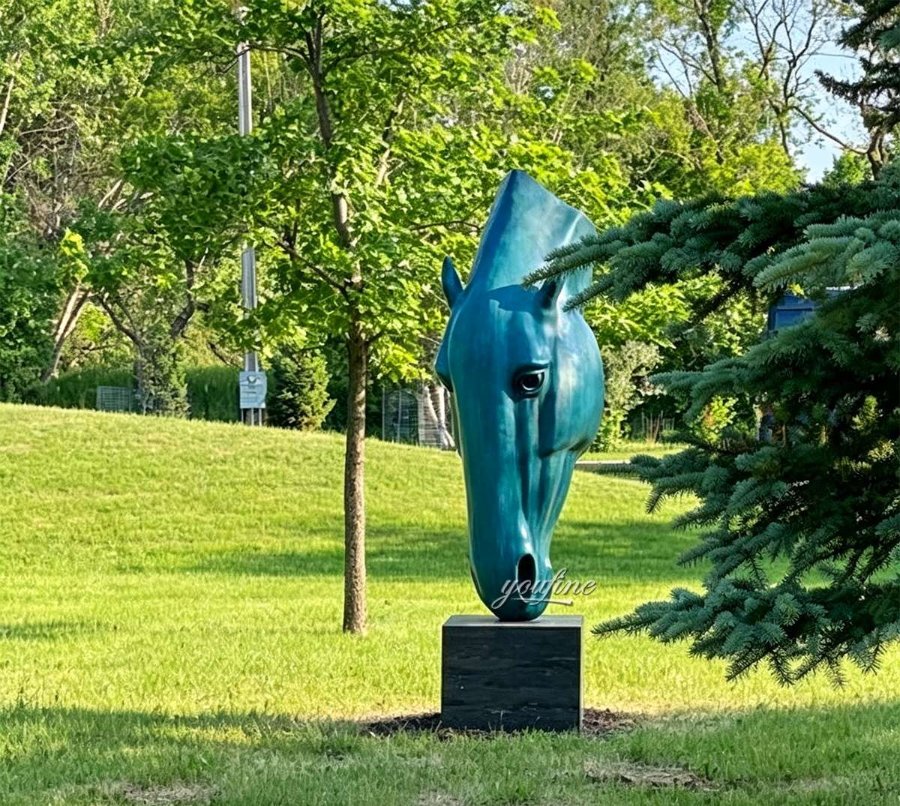 A turquoise horse head sculpture on a black pedestal in a lush green garden with trees and grass under sunlight.