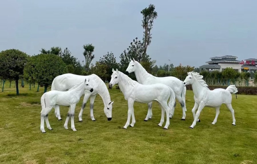 A family group of white fiberglass horse statues, including adult horses and foals, installed on a grass lawn in an outdoor park setting